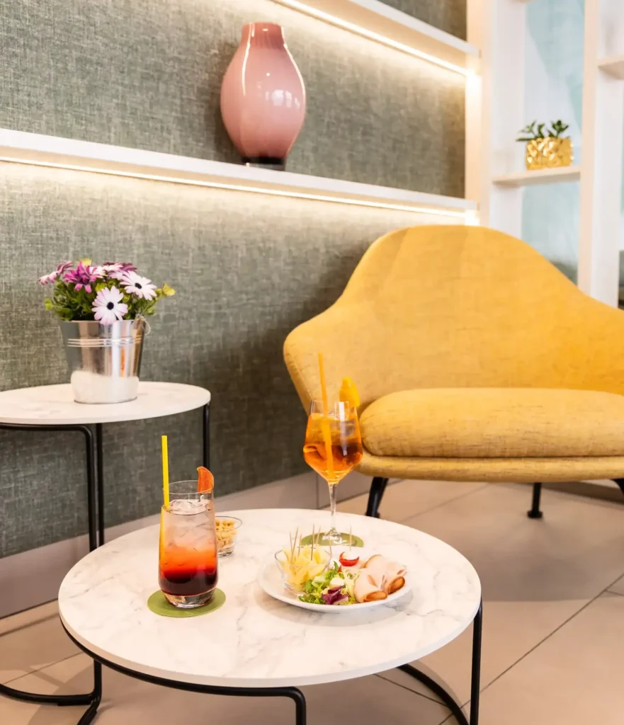 In the picture, we can see a detail of the Hall of the Hotel Luxor with a yellow armachair and a round table with two cocktaiuls and some snacks.